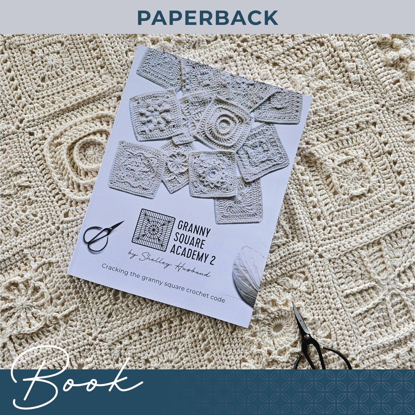 Granny Square Academy 2 front cover logo, sitting below 8 crocheted granny squares scattered over a white background. Black yarn scissors are in the bottom left corner and a ball of parchment coloured yarn sits in the bottom right. Words over the image say Paperback Book.