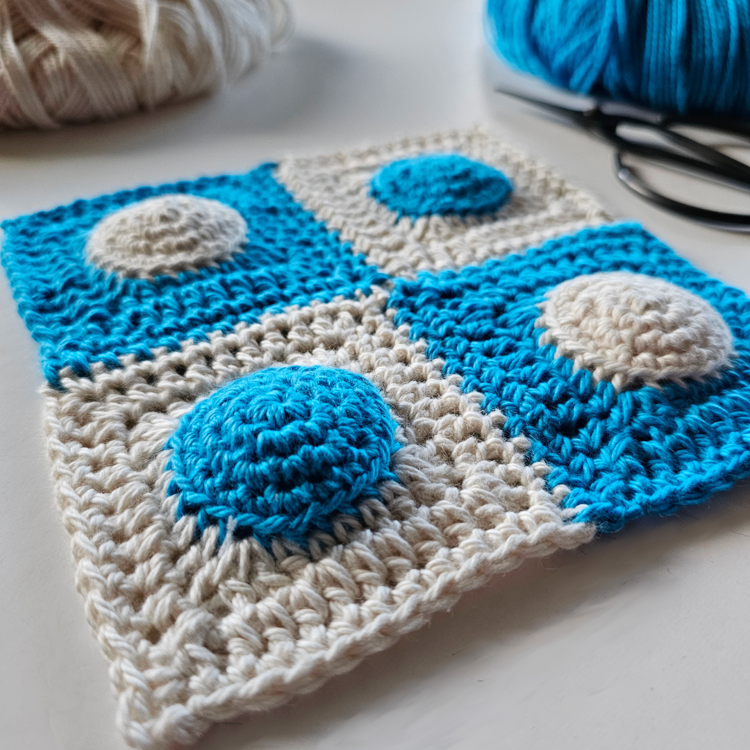 A joined 2 by 2 grid of four granny squares crocheted in blue and parchment. A half-used ball of parchment yarn sits to the top left and a half-used ball of blue sits to the top right, with a pair of black yarn scissors.
