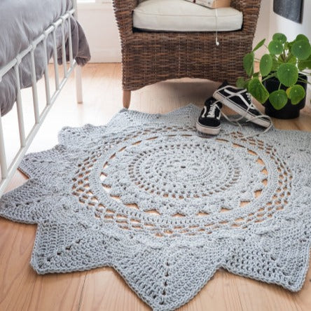 A bedroom scene with a Radiance Floor Rug by Shelley Husband in grey in the middle of the image on a slight stained hardwood floor. The rug has a circular design that it mandala like that has triangular edges all around the outside. To the left of the rug a white metal framed bed with a grey quilt cover. Behind the rug is a rattan woven, high back armchair with seat cushion and decorative cushions.