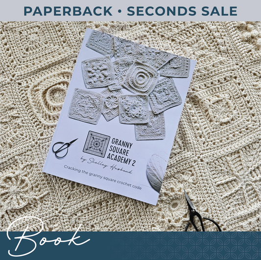 Granny Square Academy 2 front cover logo, sitting below 8 crocheted granny squares scattered over a white background. Black yarn scissors are in the bottom left corner and a ball of parchment coloured yarn sits in the bottom right. Words over the image say Paperback Book Seconds Sale!