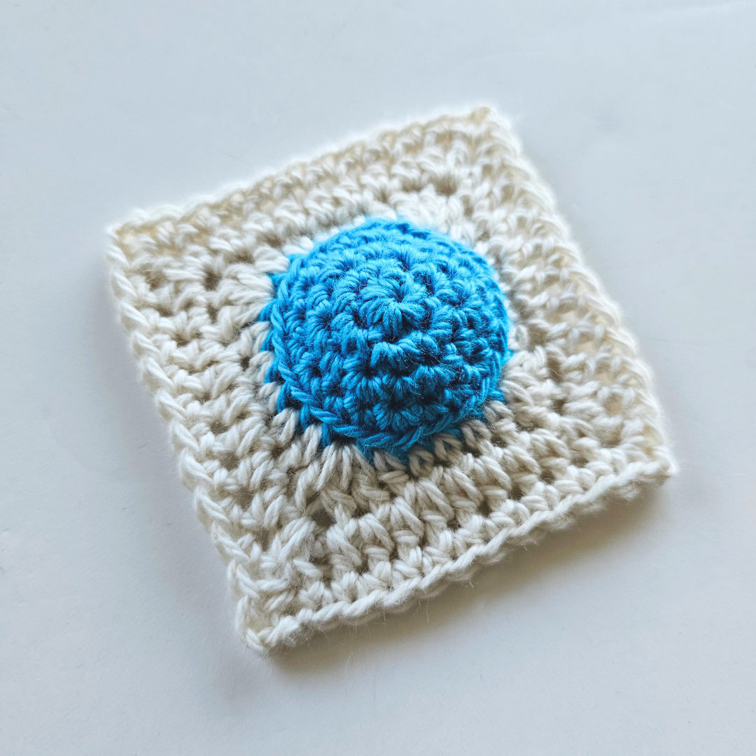 A single granny square, made up of a blue circular middle and a parchment frame to make it square.