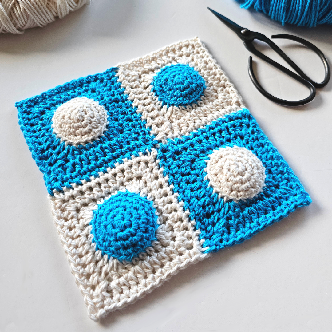 A joined 2 by 2 grid of four granny squares crocheted in blue and parchment. A half-used ball of parchment yarn sits to the top left and a half-used ball of blue sits to the top right, with a pair of black yarn scissors.