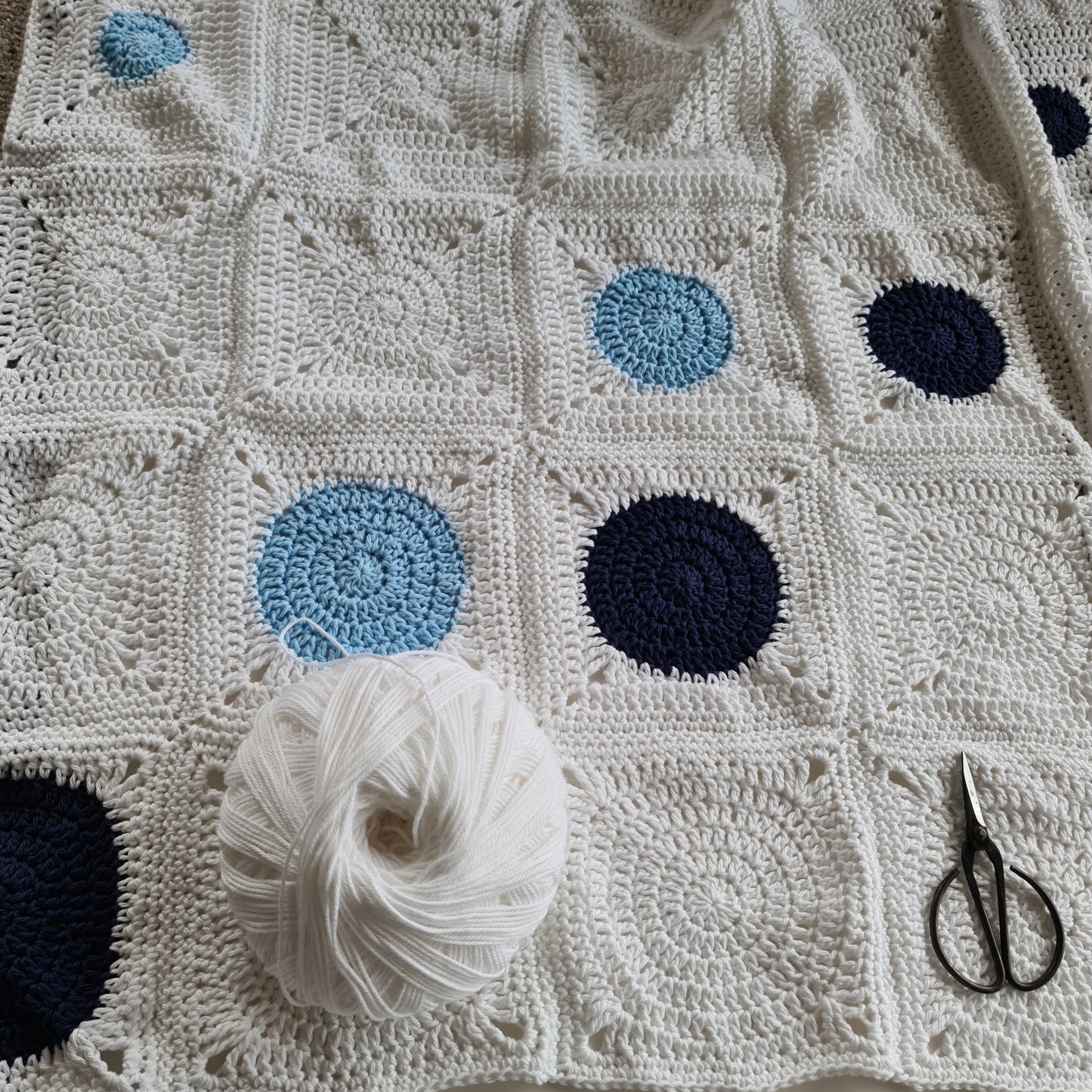 White and blues random version of the Dotty Spotty Blanket with a ball of white yarn and black scissors.