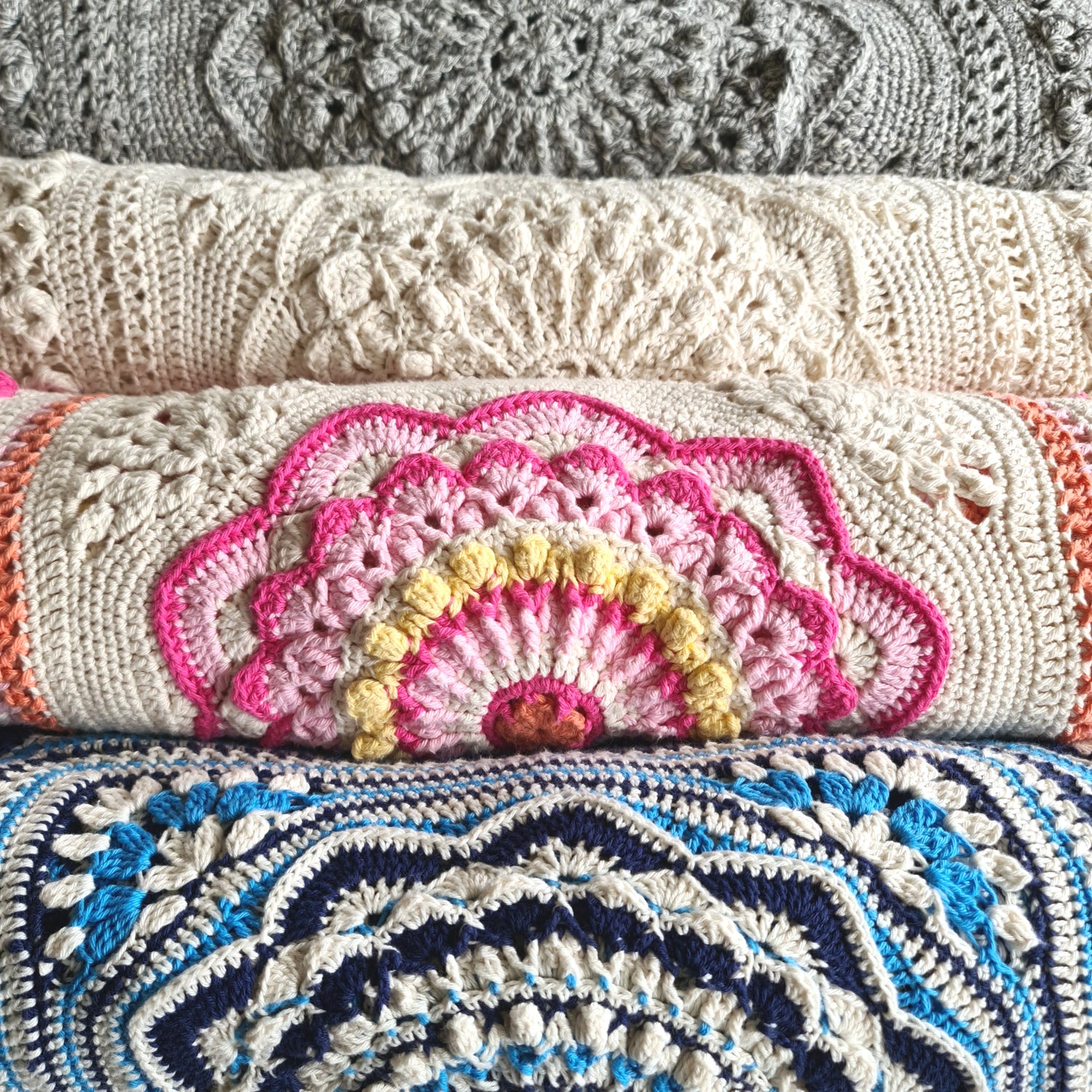 Four folded Nimue blankets showing the fold are shown horizontally. From top, a luxe mid-grey suri alpaca and merino blend blanket, a cream cotton blanket, a colourful pink and cream with hints of yellow and orange version  and a bright blue, navy and cream version at the bottom.