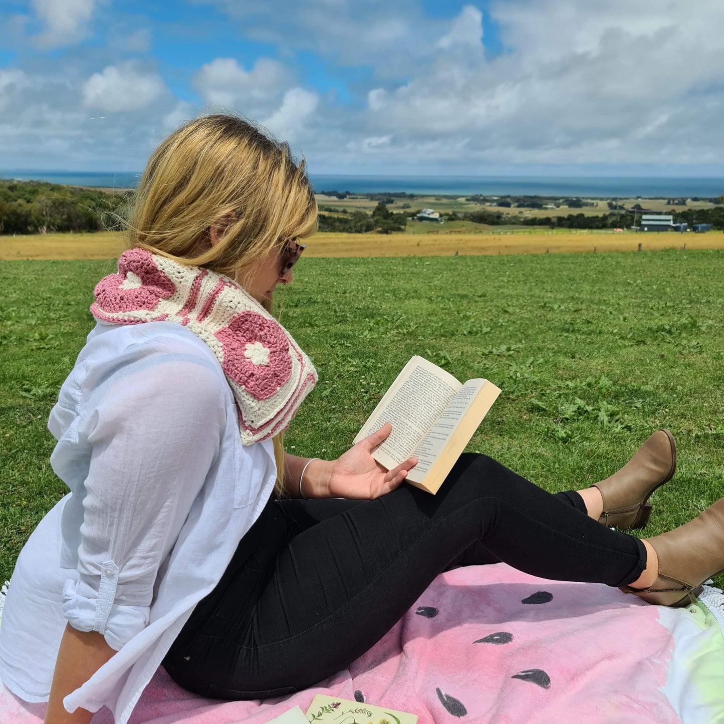 Iris Cowl in pink and cream by Shelley Husband being worn by a blonde woman reading in a paddock