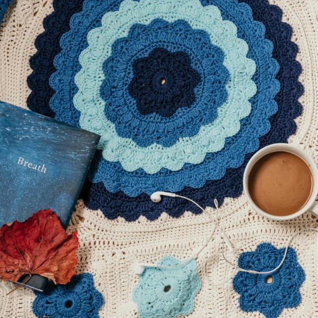Manderley Crochet Blanket by Shelley Husband with a cup of coffee, a book and headphones sitting on it
