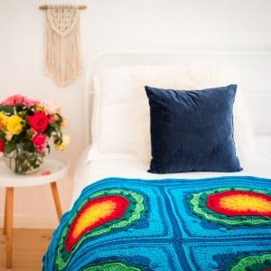 Mayan Crochet Blanket Pattern by Shelley Husband in bright colours over a bed with a navy blue cushion and a vase of roses on the sidetable
