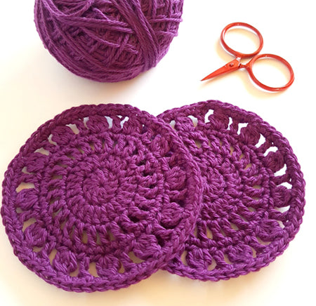 Chrissy's Coasters Pattern by Shelley Husband close up in bright purple with a ball of yarn and red yarn scissors