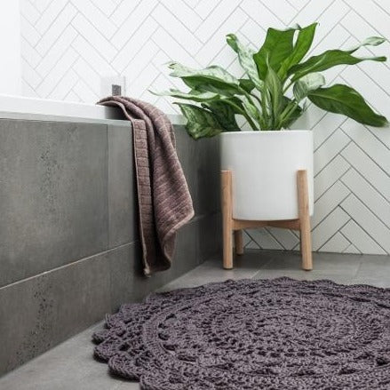 A purple Radiance Floor Rug by Shelley Husband sits on the bathroom floor next to a bath. The floor is mid grey tiles and the edge of the bath is dark grey stone tile. There is a brown bath towel hanging over the bath’s edge. A herringbone pattern of long subway tiles in with with dark grout lines the back wall. At the back edge of the bath, behind the rug, is a large white planter on a light oak stand, with a fiddle leaf like plant inside.