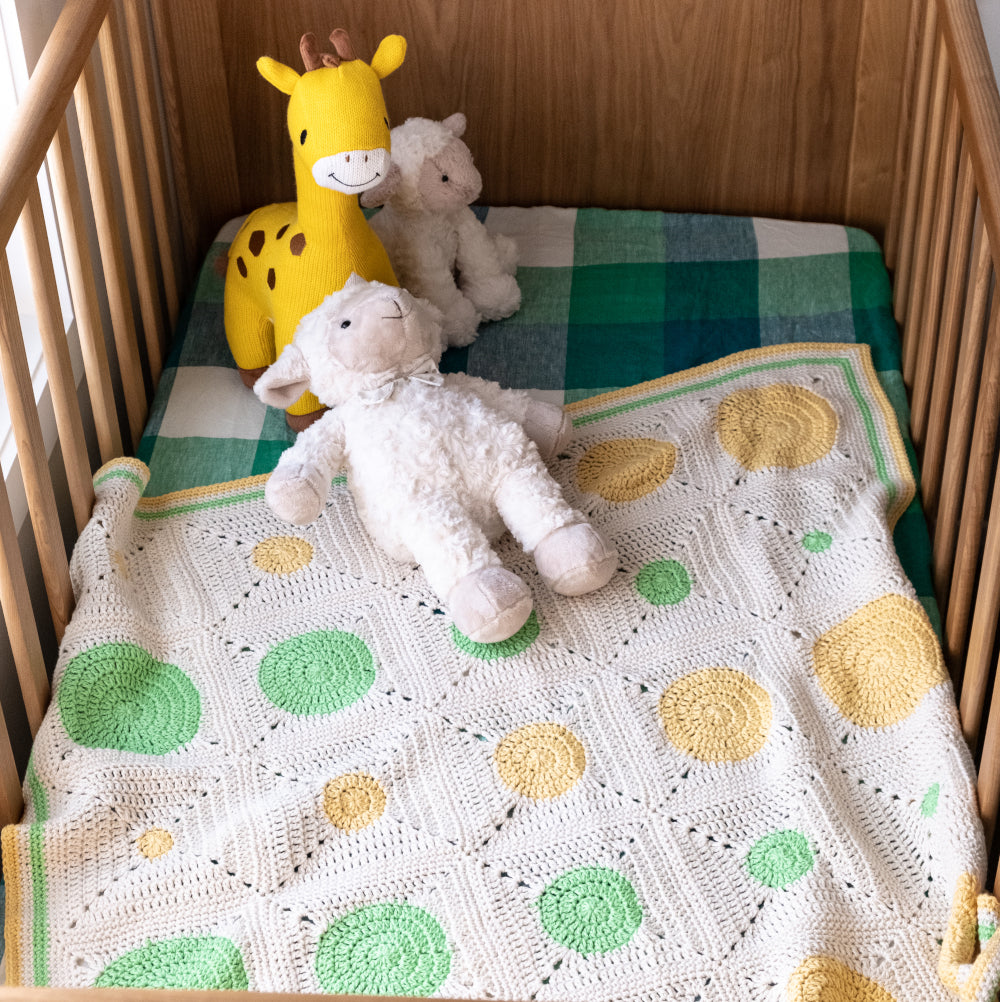 A parchment yarn Dotty Spotty blanket with bright green and pale yellow circles lays inside a natural timber cot over a buffalo check green, navy blue and white cot mattress with two lamb toys and a giraffe soft toy.