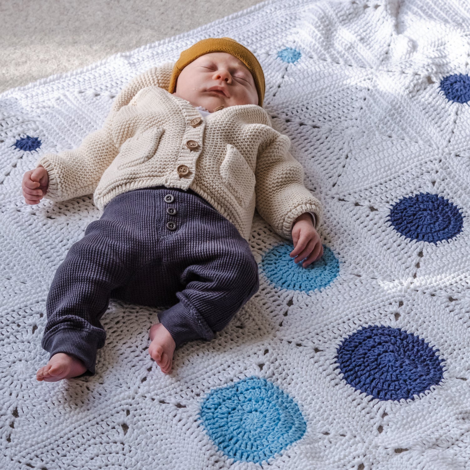 Small baby naps on their back on a white cotton Dotty Spotty Blanket with two shades of blue circles and white on white circles on a carpeted floor.