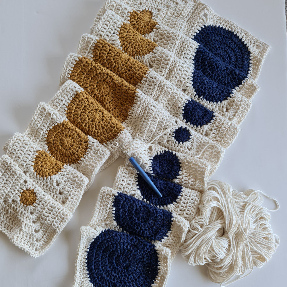 Two rows of Dotty Spotty work in progress with the left in mustard and the right in navy blue being crocheted together with parchment yarn and a light blue handled crochet hook.