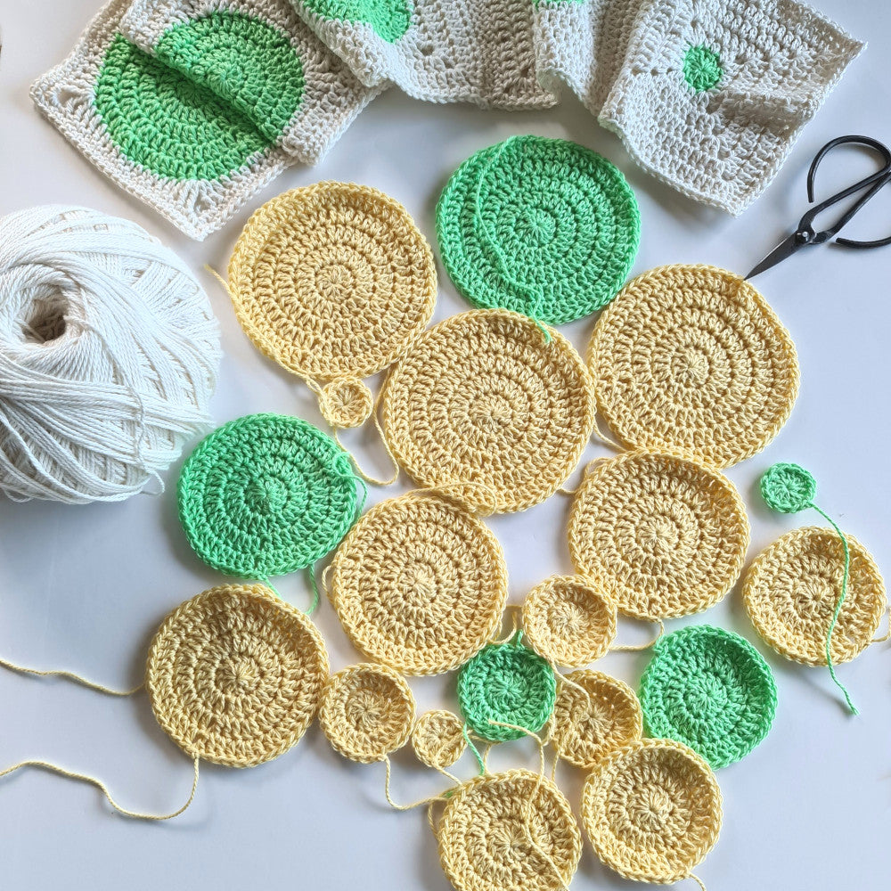 Pale yellow and bright green circles from an in progress Dotty Spotty Blanket with a ball of yarn on the left and scissors on the right.