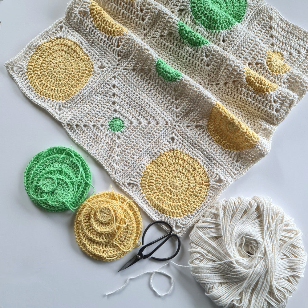 Pale yellow and bright green granny squares stitched in partially finished rows with loose circles waiting to become granny squares and parchment yarn sitting at the lower right corner.